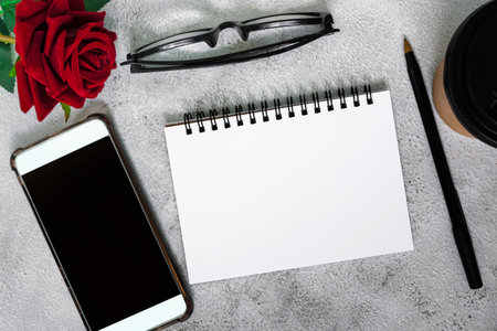 Note book with disposable coffee cup, pen, smartphone and roses on white marble table. Directly...
