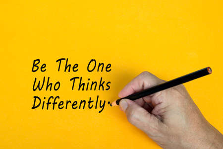 Male hand writes in black pencil the word BE THE ONE WHO THINKS DIFFERENTLY on a yellow background. Encouragement words.の写真素材