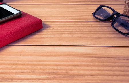 Cellphone, red note book, potted plant and reading glasses on wooden desk. Copy space.の写真素材