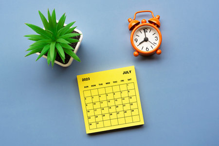 July 2023 calendar on Adhesive Note with potted plant and alarm clock set at 8 o'clock against blue...
