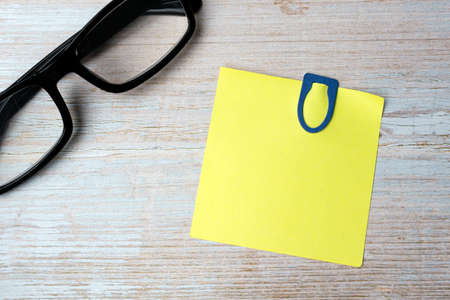 Yellow stick note with blue paperclip and reading glasses on wooden desk. Directly above. Copy space. Flat lay.の写真素材