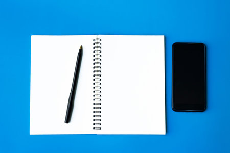 Top view of smart phone, pen and notepad on blue background. Copy space. Flat lay.