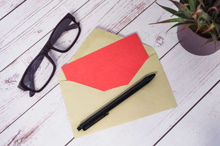 Red card mockup with envelope, pen, reading glasses and potted plant on wooden table. Copy space. Flat lay. Invitation concept.の写真素材