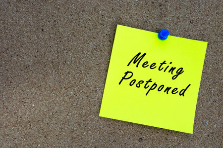 Meeting postponed on yellow stick note and pinned to a cork notice board. Reminder and business concept.の写真素材