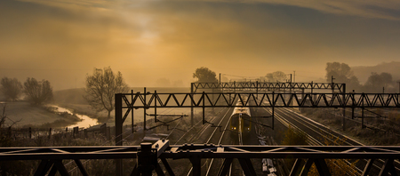 Morning express train passing along the tracks on a misty morning, with the river bathed with a golden glow from the sunriseの写真素材