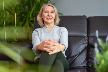 Lovely middle-aged blond woman sitting on a sofa at home looking at the cameraの写真素材
