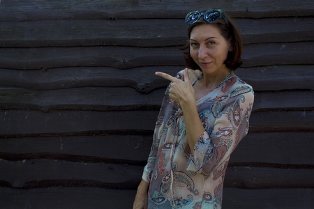 Cute woman points a finger at an empty screen area. Looks at You. Against the background of a wooden wall.の写真素材