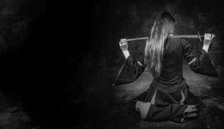 full length portrait of young long haired woman wearing a medieval costume. sitting pose with a sword over her shoulders at dark studio background with copy text to the leftの写真素材
