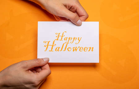 female hands holding a piece of paper with the inscription happy halloween in orange writing over orange background cardboardの写真素材