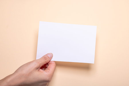 female hand holds a blank piece of paper with copy space in front of a cream colored background...