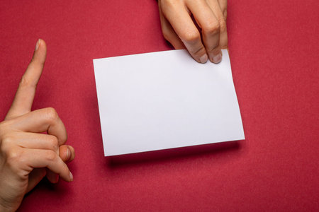 female hand holds a blank piece of paper with copy space in front of a dark red background cardboard...
