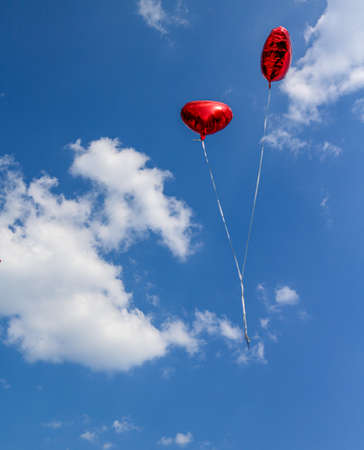 two red balloons in heart shape in front of a blue summer sky with white fair weather cloudsの写真素材