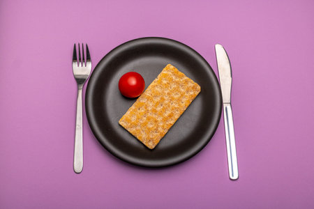 A single tomato and a slice of crispbread on a plain black plate and cutlery with signs of use against a purple backgroundの写真素材