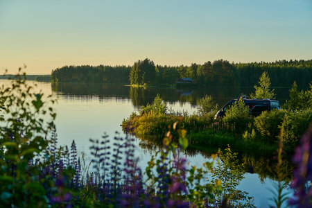Camper van parked at a lake at sunset or sunrise at spring or early summer evening with clear sky background and colorful flowers along the shore of a very calm and flat dramatic lake with water reflection in Finland at midsummer.の写真素材
