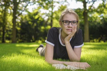A young woman with glasses lies on the grass in the park and looks happy at the cameraの写真素材