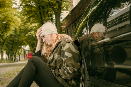 A young woman with a head injury leans against a car and looks painfullyの写真素材