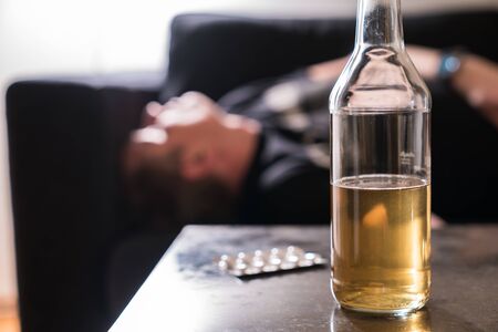 a drunk man with alcohol bottle and pills on the table sleeping on couchの写真素材