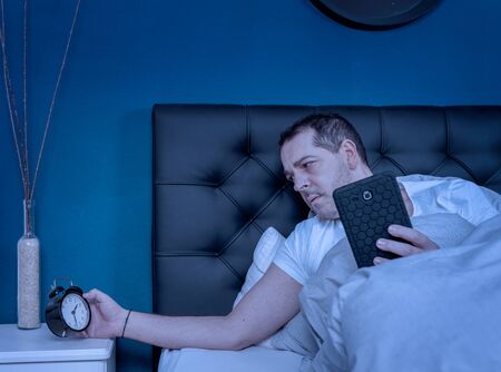 a man working on his smartphone at night looks at his alarm clock and cannot sleepの写真素材