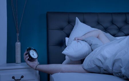 a sleepless man hold his pillow and alarm clock at nightの写真素材
