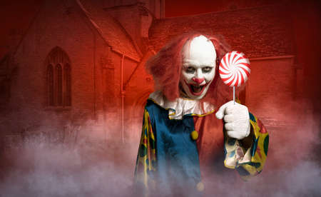 a scary clown in front of a old church tries to lure with a lollyの写真素材