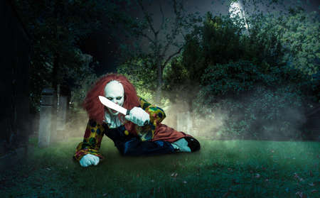 a scary clown lies with a knife on a cemetery threatens the viewerの写真素材
