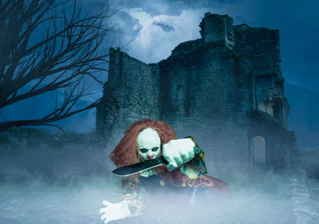 an evil clown with a knife lies in front of an old castleの写真素材