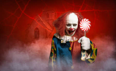 an eerie clown in front of a red background tries to lure with a lollyの写真素材