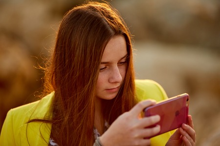 Close up of beautiful red-haired girl uses a cell phone sitting in social network on the shore and seaの写真素材
