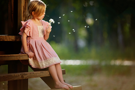 Happy small girl blowing dandelion flower outdoors. Girl having fun in spring park. Blurred background in sunsetの写真素材