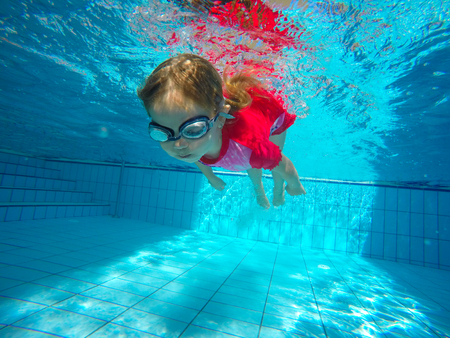 Funny portrait of baby girl swimming and diving in blue pool with fun - jump from poolside deep down underwater with splashes. Family lifestyle and summer children water sports activity with parents.の写真素材