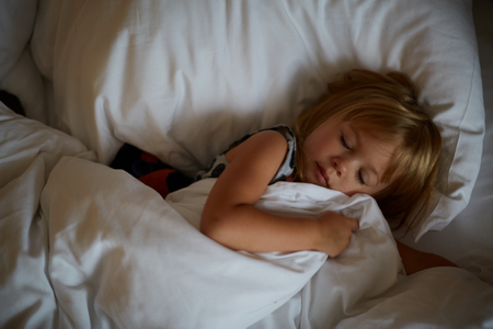 View of lovely girl with long light hair sleeps after reading fairy tale, lies in comfortable white bed, has pleasant dreams, enjoys fairytale. Children and relaxation conceptの写真素材