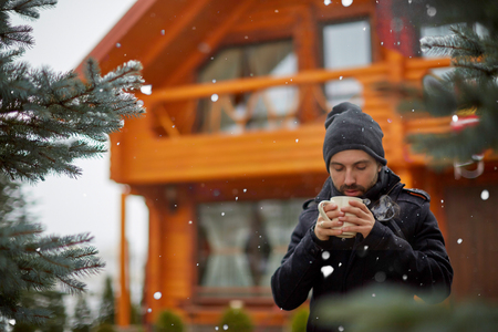 Young man in outwear having cup of warming drink standing outdoors of cottage in snowfallの写真素材