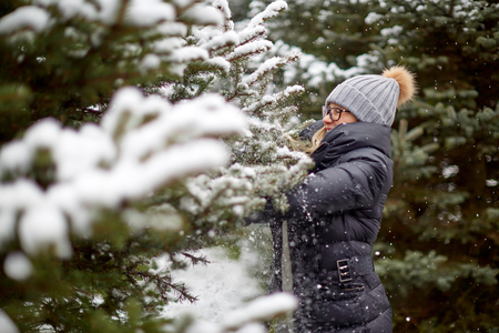 Side view of pretty young female in warm clothes smiling and touching conifer tree covered with snow on amazing winter dayの写真素材