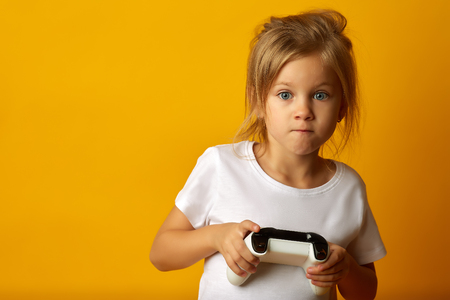 Little absorbed girl in white t-shirt holding game pad playing video game on yellow backgroundの写真素材