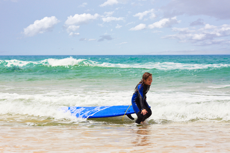 Side view of sweet girl in swimsuit smiling and pulling blue surfboard out of waving sea while having fun on sunny dayの写真素材