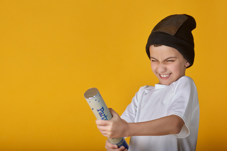Playful little boy in white t-shirt and hat exploding party popper standing on yellow backdropの写真素材