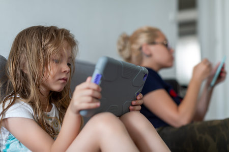 Girls are sitting on sofa and reading. Woman holds tablet while girl has phone in hands
