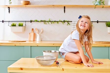Happy little girl with blond hair cheerfully smiling and looking up while sitting on knees on wooden table near utensils in kitchenの写真素材