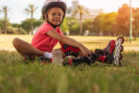 Cute little blonde girl sitting on green grass and putting on roller skatesの写真素材