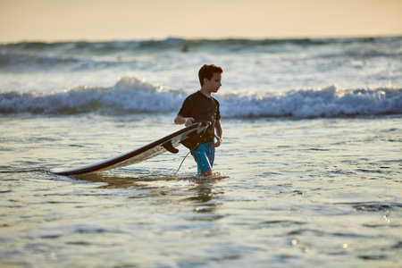 Side view of fearless kid floating surfboard at ocean with waves on sunny eveningの写真素材