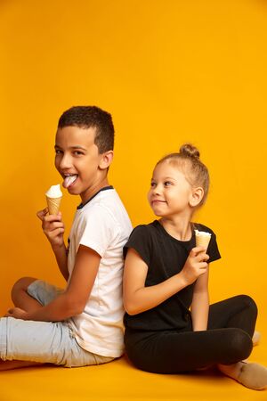funny kids eat vanilla ice cream in a waffle cone on a yellow background, joyful brother and sisterの写真素材