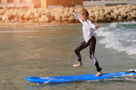 Summer vacation with child. Happy baby girl young surfer ride on surfboard with fun on sea waves. Active family lifestyle, kids outdoor water sport lessons and swimming activity in surf campの写真素材