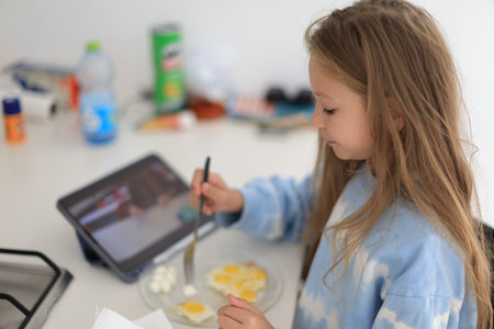 Positive caucasian little girl having breakfast and looking at tablet at homeの写真素材