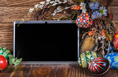 Easter composition. Holiday wreath with colored eggs and tablet pc with blank screen on a wooden table. Easter themeの写真素材