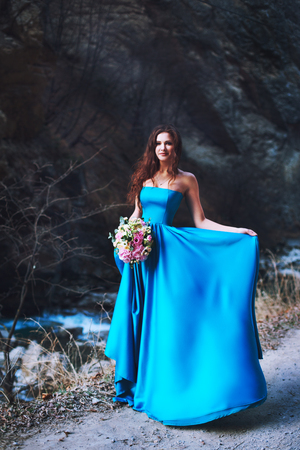 The brunette at the foot of the mountain stands in a blue dress with a bouquet of flowersの写真素材