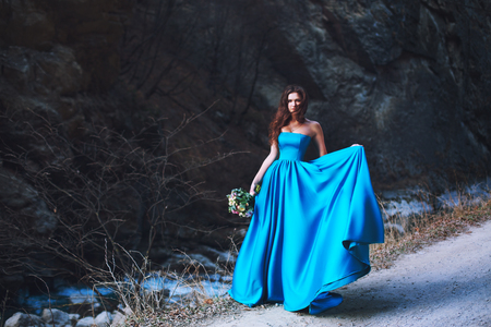 A beautiful girl with a wedding bouquet and in a blue dress is standing at the foot of the mountainの写真素材