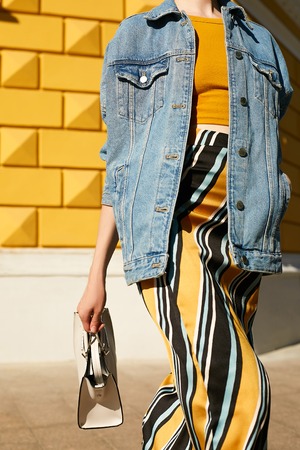 Fashionable girl in a denim jacket, yellow top and in a skirt with an ornament in a hurry, waving a white square bag. Street lifestyle.の写真素材