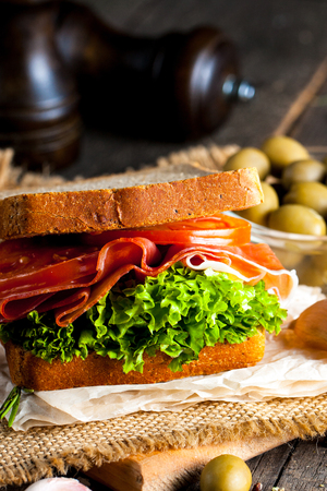 Close-up photo of a club sandwich. Sandwich with meat, prosciutto, salami, salad, vegetables, lettuce, tomato, onion and mustard on a fresh sliced rye bread on wooden background. Olives background.の写真素材