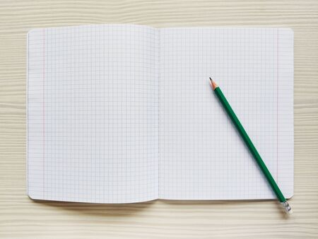 White school notebook in a cage is on a wooden table. Green pencil rests on a checkered exercise book without notes. Empty notepad is on a desk. Square textureの写真素材