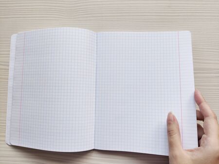 White open school checkered exercise book in a cage is on a wooden table. White woman opens a notebook without notes. The right hand of a white European woman holding a empty notepad with squareの写真素材